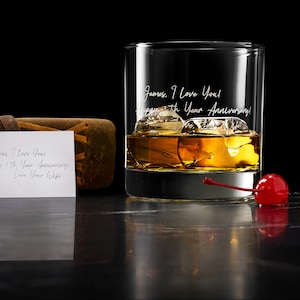 May include: A clear glass tumbler filled with amber liquid and ice cubes, featuring the engraved text: James, I Love You! Happy 5th Year Anniversary! Love, Your Wife! A bright red maraschino cherry sits nearby, alongside a small white note card.