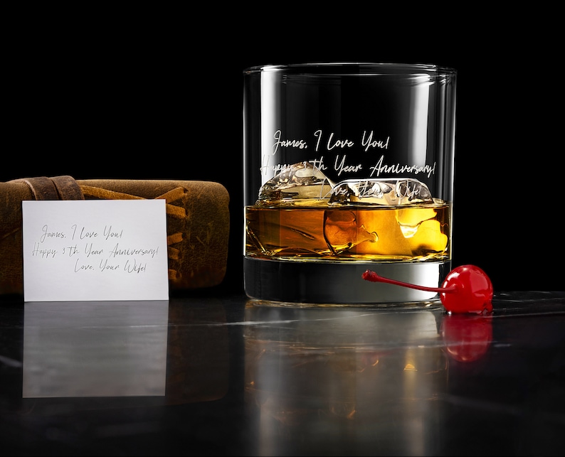 May include: A clear glass tumbler filled with amber liquid and ice cubes, featuring the engraved text: James, I Love You! Happy 5th Year Anniversary! Love, Your Wife! A bright red maraschino cherry sits nearby, alongside a small white note card.