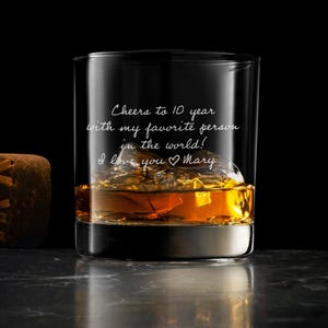 May include: A clear glass tumbler filled with amber liquid and ice cubes, featuring white engraved text that reads: Cheers to 10 year with my favorite person in the world! I love you Mary. The glass has a thick base and sits next to a brown leather item.
