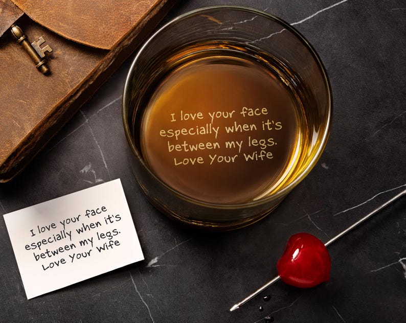 May include: A clear glass tumbler containing amber liquid with the etched text: I love your face especially when it's between my legs. Love Your Wife. A matching white card with the same text, a red cherry on a metal pick, and a brown leather item.