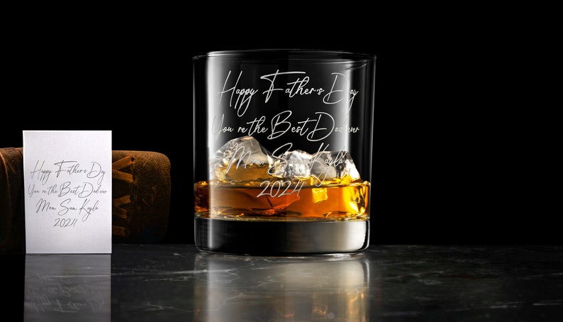 May include: A clear glass tumbler filled with amber liquid and ice cubes, featuring white engraved text that reads Happy Father's Day, You're the Best Dad ever, Love, Son, Kayla, 2024. A matching white card with the same text is visible.
