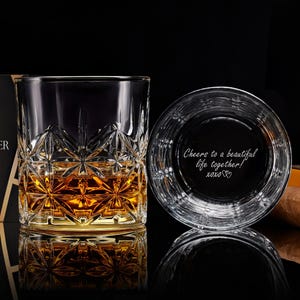 May include: A clear glass tumbler with a faceted star pattern containing amber liquid, next to a second glass showing the engraved text "Cheers to a beautiful life together! xoxo". A brown cigar and a black gift box are also visible.
