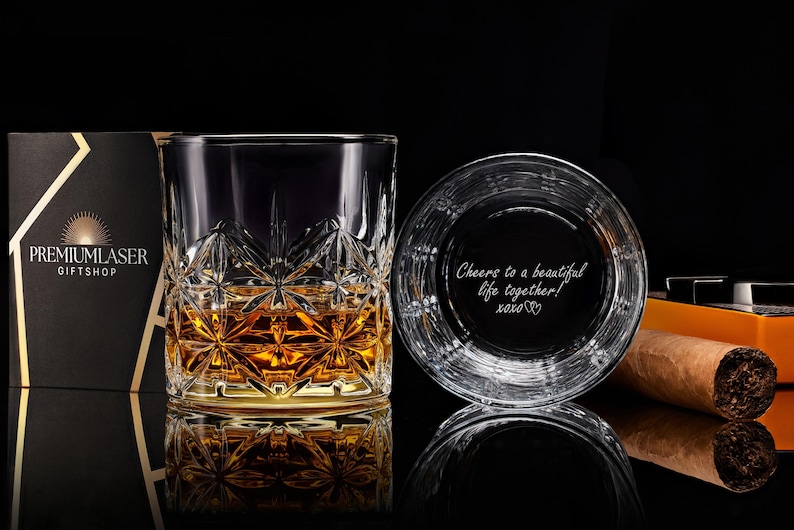 May include: A clear glass tumbler with a faceted star pattern containing amber liquid, next to a second glass showing the engraved text "Cheers to a beautiful life together! xoxo". A brown cigar and a black gift box are also visible.