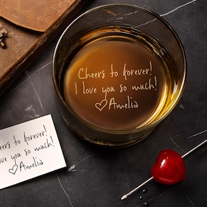 May include: A clear glass tumbler filled with amber liquid, featuring the engraved text "Cheers to forever! I love you so much! ♡Amelia". Beside it lies a white card with the same handwritten message and a metal cocktail pick with a red cherry.