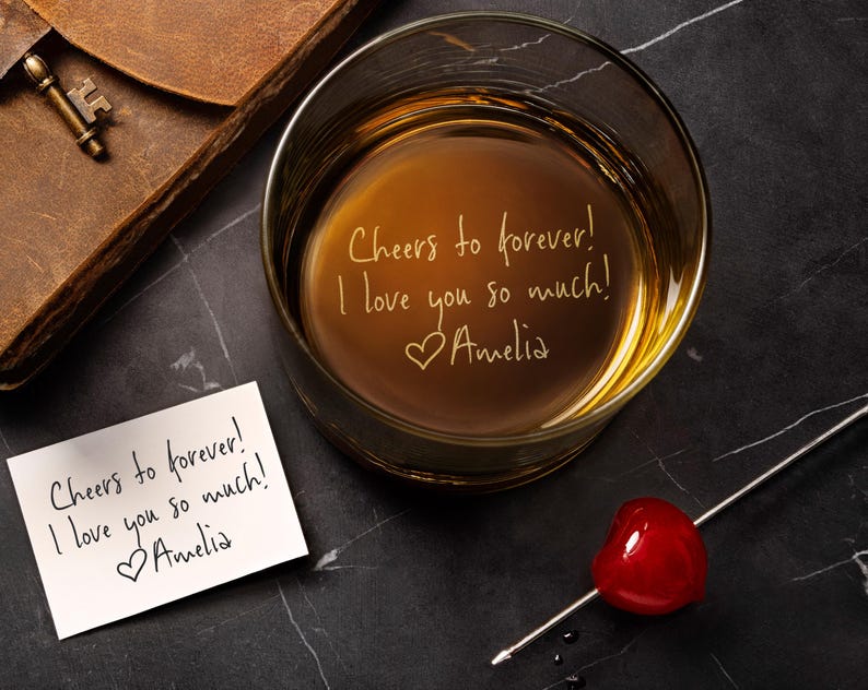 May include: A clear glass tumbler filled with amber liquid, featuring the engraved text "Cheers to forever! I love you so much! ♡Amelia". Beside it lies a white card with the same handwritten message and a metal cocktail pick with a red cherry.
