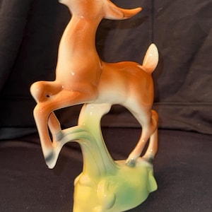 May include: A ceramic figurine of a brown deer standing on its hind legs with its head tilted back and mouth open. The deer is standing on a green and yellow base.