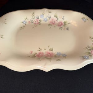 May include: Cream-colored, oval-shaped serving platter with a scalloped edge. The platter is decorated with pink and blue floral patterns and green leaves. A decorative piece for serving food.
