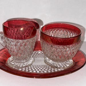 May include: A vintage ruby red glass creamer and sugar bowl set with a diamond pattern, resting on a matching oval tray. The rims of the creamer, sugar bowl, and tray are a vibrant red. The set is likely from the mid-20th century.