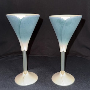 May include: Two elegant, tulip-shaped wine glasses. The bowls are a muted blue with a white interior, transitioning to a clear stem and a cream-colored base. The design is reminiscent of a flower, adding a touch of sophistication.