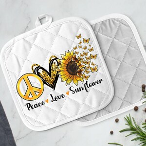 May include: White quilted pot holders with a peace sign, heart, and sunflower design. The text reads "Peace Love Sunflower".