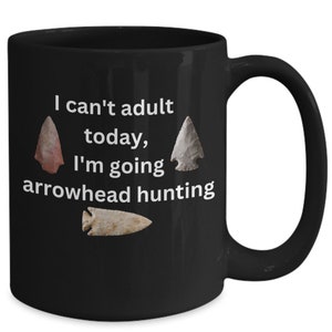 May include: Black coffee mug with white text that reads "I can't adult today, I'm going arrowhead hunting". Three arrowheads are pictured on the mug.