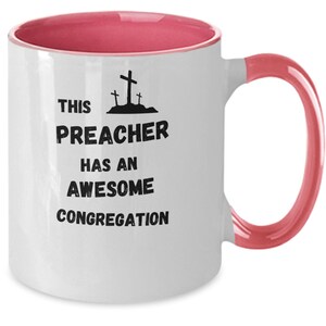May include: White ceramic coffee mug with a pink rim and handle. The mug has black text that reads "This Preacher Has An Awesome Congregation" with a graphic of three crosses.