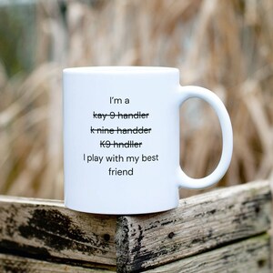 May include: White ceramic coffee mug with black text that reads "I'm a kay 9 handler k nine handder K9 hndiler I play with my best friend"