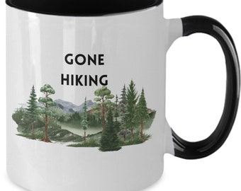 May include: White ceramic coffee mug with a black interior and handle. The mug features a black and white graphic of a forest scene with the text "Gone Hiking".