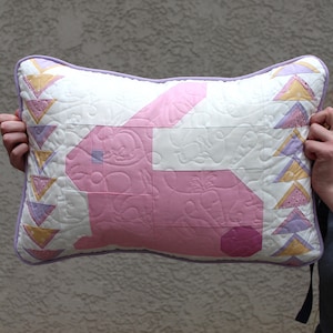 This shows the front of the pillow. There is a light pink side profile of a bunny with a light purple eye and dark pink tail. The background of the pillow is white. On the sides there are yellow, pink and purple triangles.