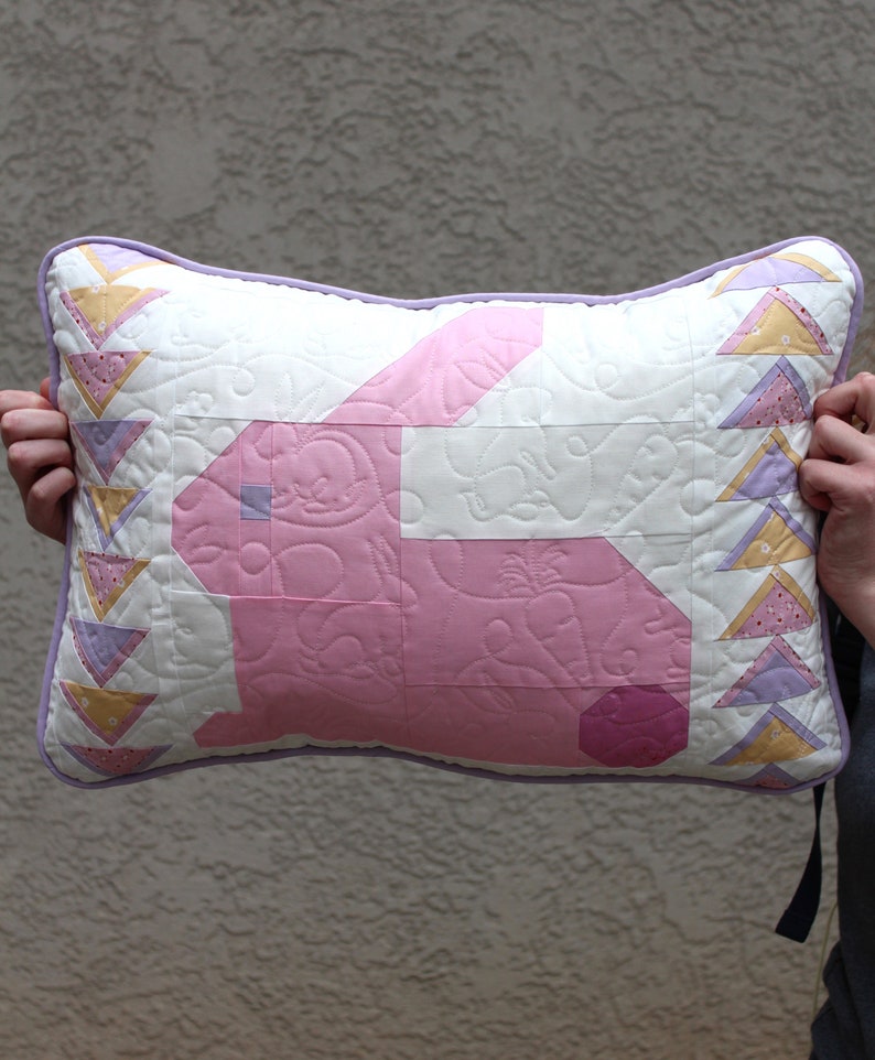 This shows the front of the pillow. There is a light pink side profile of a bunny with a light purple eye and dark pink tail. The background of the pillow is white. On the sides there are yellow, pink and purple triangles.