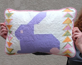 May include: A purple bunny-shaped pillow with a white background and a pink border. The pillow is decorated with a patchwork of colorful triangles.