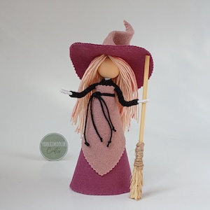 Peut inclure: Une poupée sorcière en feutrine avec un chapeau pointu rose foncé et une robe assortie dotée d'un tablier rose clair. La poupée a de longs cheveux en laine rose clair, des bras noirs et un visage beige. Elle tient un balai en bois. Logo YOOLEEMOOLEE Crafts.