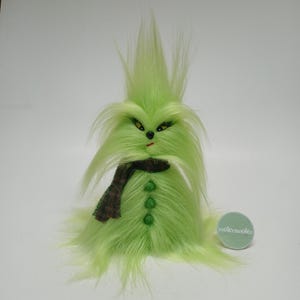 May include: A fuzzy lime green creature figurine with long, spiky hair, yellow eyes, a small black nose, and a red mouth. It wears a brown plaid scarf and features three green buttons down the front. A round tag with the text yooleemoolee is visible.