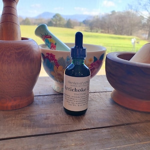 May include: A dark green glass bottle with a dropper, labeled "Garden of Pepper" and "Artichoke Leaf Tincture". The bottle is sitting on a wooden surface with two wooden mortars and a blue and white ceramic mortar and pestle.