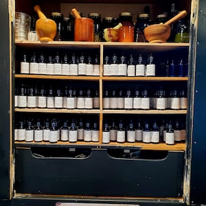 May include: A wooden cabinet with shelves filled with small glass bottles. The cabinet is labelled "APOTHECARY" and has a drawer below the shelves. The shelves are filled with various herbs and spices.