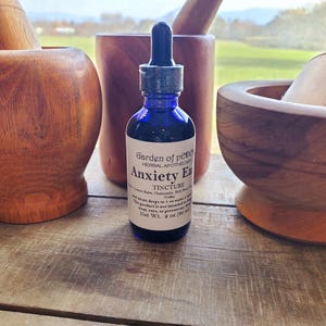Puede incluir: Una botella de vidrio azul con un gotero etiquetada como "Garden of Peace Herbal Apothecary Anxiety Ease Tincture" con una lista de ingredientes que incluyen manzanilla, albahaca sagrada y vodka. La botella est&aacute; sobre una superficie de madera con dos morteros y mazos de madera.