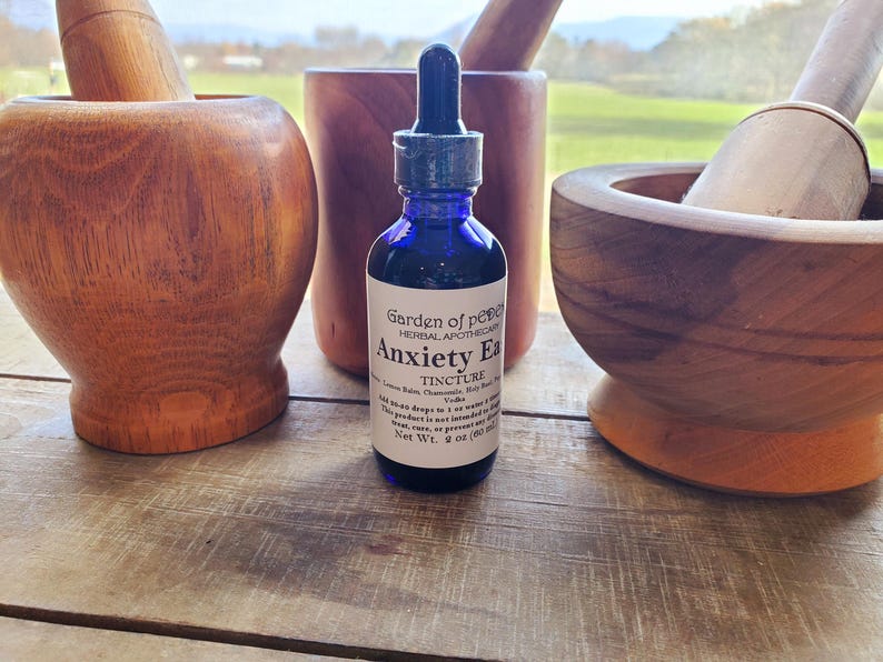 Puede incluir: Una botella de vidrio azul con un gotero etiquetada como "Garden of Peace Herbal Apothecary Anxiety Ease Tincture" con una lista de ingredientes que incluyen manzanilla, albahaca sagrada y vodka. La botella est&aacute; sobre una superficie de madera con dos morteros y mazos de madera.