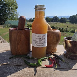 May include: A clear glass bottle of Garden of Eden Herbal Apothecary Fire Cider with a white label, flanked by two wooden mortars and pestles and a small stone mortar and pestle, surrounded by several fresh green and red chili peppers.