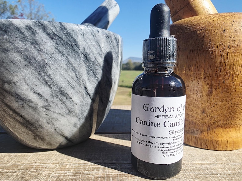 May include: A dark glass bottle with a dropper, labelled "Garden of Herbal Apothecary Canine Candid". The bottle is next to a large grey and white marble mortar and a wooden pestle. The background includes a field and blue sky.