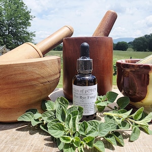 May include: A wooden mortar and pestle set with a small bottle of oil and fresh green herbs. The oil bottle has a dropper and a label that reads "Bye Bye Fungi". The background includes a blurred outdoor scene.