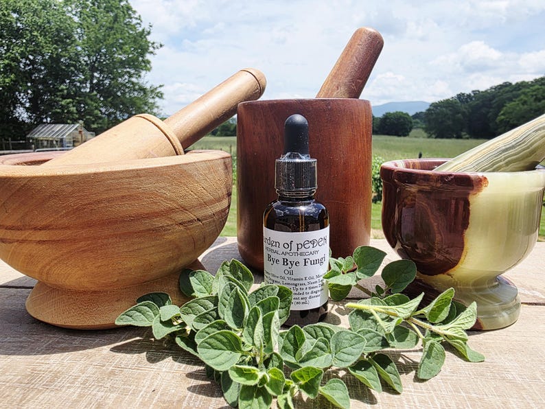 May include: A wooden mortar and pestle set with a small bottle of oil and fresh green herbs. The oil bottle has a dropper and a label that reads "Bye Bye Fungi". The background includes a blurred outdoor scene.