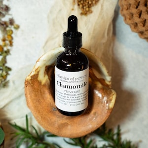May include: A small brown glass bottle with a dropper top, labeled "Garden of pEDEN HERBAL APOTHECARY Chamomile TINCTURE" and "Net Wt. 2 oz (60 ml)". The bottle is sitting on a small, brown ceramic dish.