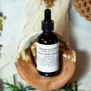May include: A dark brown glass bottle with a black dropper top, labeled "Garden of pеден Herbal Apothecary Feverfew with Chamomile Tincture". The bottle is sitting on a brown and white ceramic dish. The label also states "Net Wt. 2 oz (60 ml)".