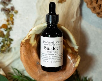 Puede incluir: Una botella de vidrio oscuro con un gotero, etiquetada "Garden of peDeN Herbal Apothecary Burdock Tincture". La botella está sentada en un plato de cerámica marrón. Los ingredientes se enumeran como Bardana, Vodka de 50 grados. La botella es de 2 oz (60 ml).