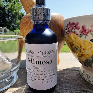 May include: A dark blue glass bottle labeled "Garden of Eden Herbal Apothecary Mimosa Tincture" with a white label. The bottle is next to a mortar and pestle and a floral patterned bowl. The bottle contains 2 oz (60 mL).