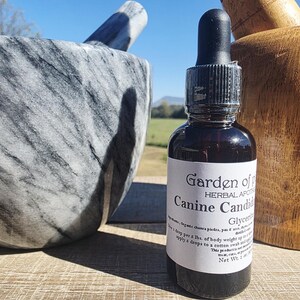 May include: A dark glass bottle with a dropper, labelled "Garden of Herbal Apothecary Canine Candida". A grey and white marble mortar and pestle are in the background, along with a wooden mortar. The bottle contains a dark liquid.