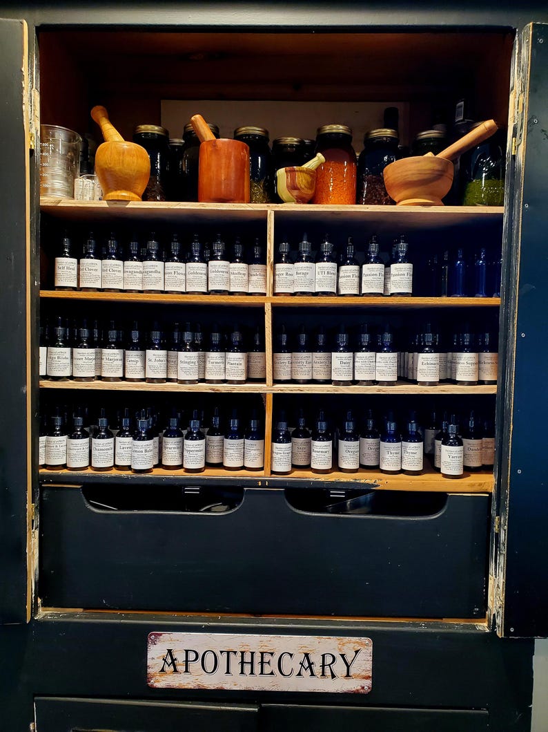 Peut inclure: Armoire d'apothicaire noire avec des &eacute;tag&egrave;res en bois, remplie de petites bouteilles en verre, de bocaux et de mortiers et pilons en bois. Les bouteilles ont des &eacute;tiquettes blanches. Le panneau en bas indique "APOTHECARY".