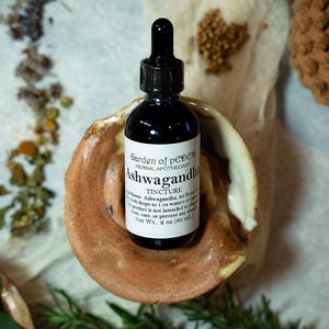 May include: A dark brown glass bottle with a dropper top, labelled "Garden of peDEN Herbal Apothecary Ashwagandha Tincture". The bottle is sitting on a small, brown ceramic dish. The label also states "Ingredients: Ashwagandha, 50 Proof Vodka. 20-40 drops to 1 oz water-1-3 times daily. This product is not intended to diagnose, treat, cure, or prevent any disease. Net Wt. 2 oz (60 ml)"
