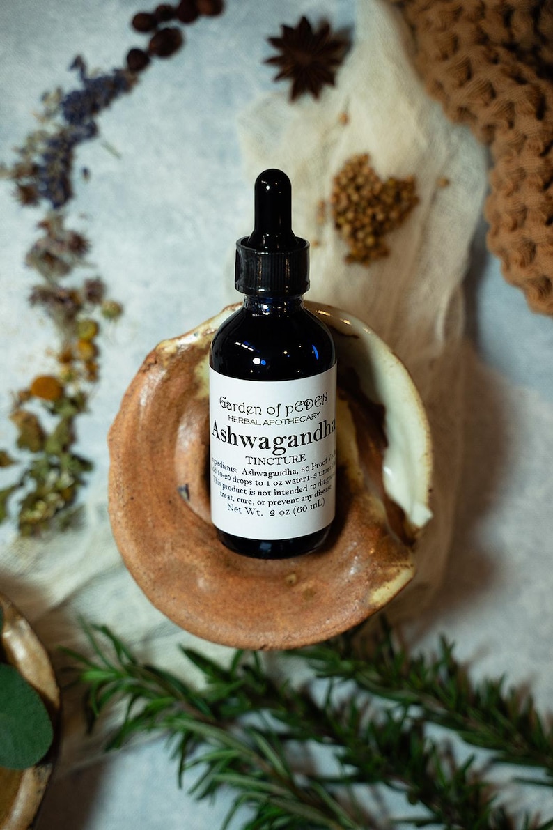 May include: A dark brown glass bottle with a dropper top, labelled "Garden of peDEN Herbal Apothecary Ashwagandha Tincture". The bottle is sitting on a small, brown ceramic dish. The label also states "Ingredients: Ashwagandha, 50 Proof Vodka. 20-40 drops to 1 oz water-1-3 times daily. This product is not intended to diagnose, treat, cure, or prevent any disease. Net Wt. 2 oz (60 ml)"
