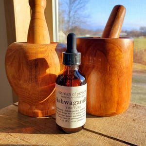 May include: A brown glass bottle with a dropper top and a white label that reads "Garden of Pepper Herbal Apothecary Ashwagandha Tincture". The bottle is sitting on a wooden surface next to a wooden mortar and pestle.