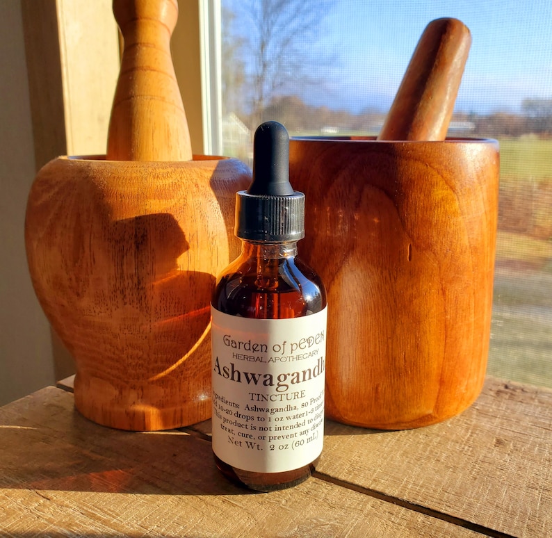 May include: A brown glass bottle with a dropper top and a white label that reads "Garden of Pepper Herbal Apothecary Ashwagandha Tincture". The bottle is sitting on a wooden surface next to a wooden mortar and pestle.