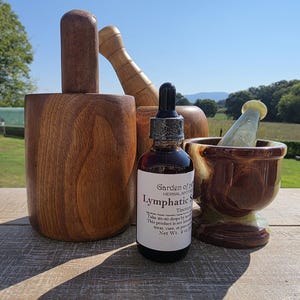 May include: A dark amber glass dropper bottle labeled Garden of Life Herbal Apothecary Lymphatic Support Tincture, positioned between a large wooden mortar and pestle set and a smaller polished brown and green stone mortar and pestle.