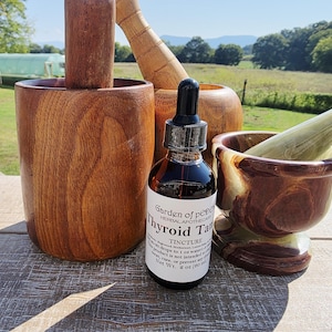 May include: A dark amber glass tincture bottle with a white label reading Garden of Peden Thyroid Tincture, positioned between a large wooden mortar and pestle set and a smaller polished brown and cream stone mortar and pestle set.