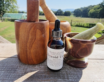 Puede incluir: Un frasco de tintura de vidrio ámbar oscuro con una etiqueta blanca que dice Garden of Peden Thyroid Tincture, colocado entre un mortero de madera grande con mano y un mortero de piedra pulida marrón y crema con mano.