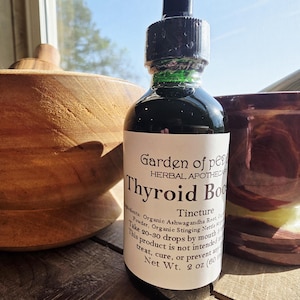May include: A dark glass bottle of "Thyroid Boost" tincture from "Garden of per Herbal Apothecary" with a black dropper. The label lists ingredients and instructions. A wooden bowl and a mortar and pestle are in the background.