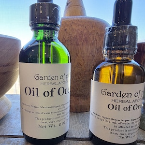 May include: Two amber glass bottles with dropper tops, labeled "Garden of p HERBAL APOTH Oil of Ore" and containing a clear liquid. The bottles are sitting on a wooden surface.