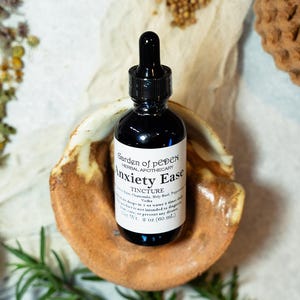 May include: A 2 ounce bottle of Anxiety Ease tincture, a herbal remedy for anxiety, made with chamomile, holy basil, and peppermint. The bottle is labeled with the name of the product and the ingredients. The bottle is sitting on a ceramic dish.