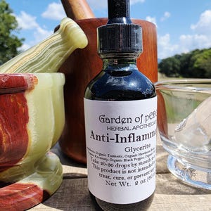 May include: A dark glass bottle with a black dropper, labeled "Garden of Peace Herbal Apothecary Anti-Inflamm Glycerite." The bottle is surrounded by a wooden mortar and pestle, and a marble mortar and pestle.