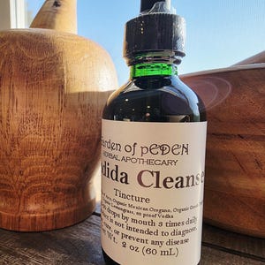Peut inclure: Une bouteille en verre fonc&eacute; de teinture "Dida Cleanse" de Garden of Peden Herbal Apothecary. L'&eacute;tiquette comprend les ingr&eacute;dients et les instructions d'utilisation. La bouteille est de 60 ml. Des objets d&eacute;coratifs en bois sont en arri&egrave;re-plan.