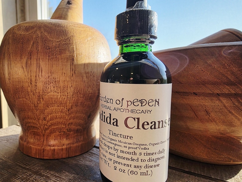 Peut inclure: Une bouteille en verre fonc&eacute; de teinture "Dida Cleanse" de Garden of Peden Herbal Apothecary. L'&eacute;tiquette comprend les ingr&eacute;dients et les instructions d'utilisation. La bouteille est de 60 ml. Des objets d&eacute;coratifs en bois sont en arri&egrave;re-plan.